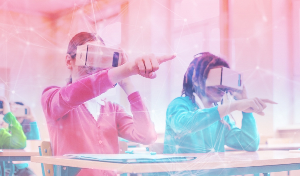 A white youth with long blonde hair in a ponytail wearing a red shirt sits next to a brown youth with dark locs in a teal shirt at a classroom table. Both youth point in front of their bodies with their right hands while holding cardboard VR headsets to their eyes with their left hands.