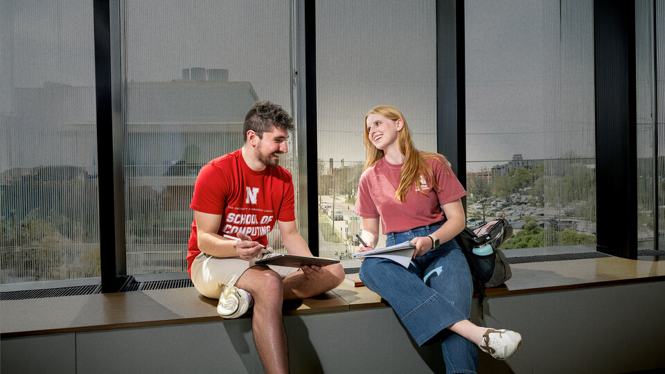 Two students talking and studying by large sunlit windows, smiling and holding notebooks.