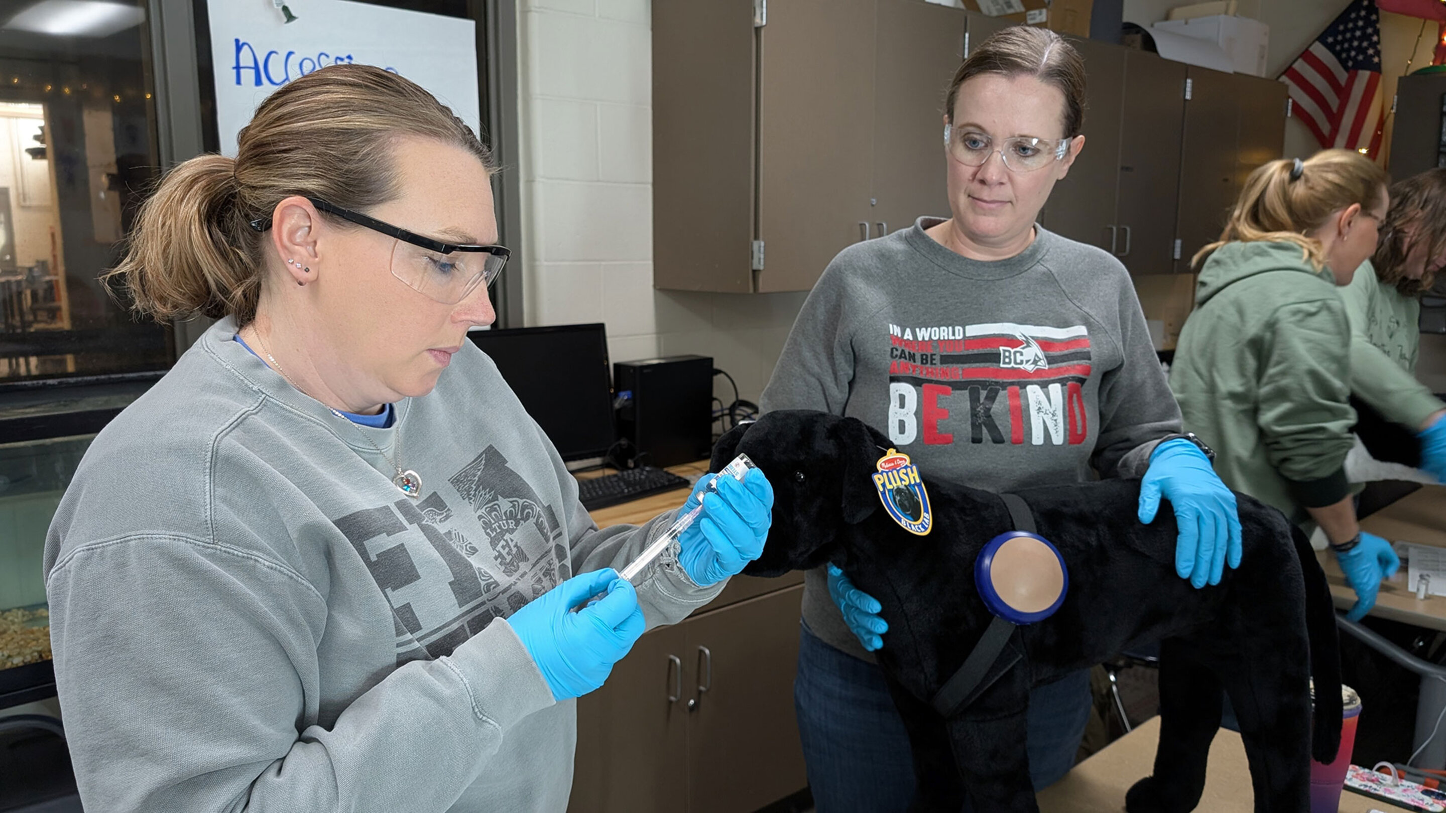 Two women in lab setting practicing medical procedures on a black dog mannequin wearing police patches.