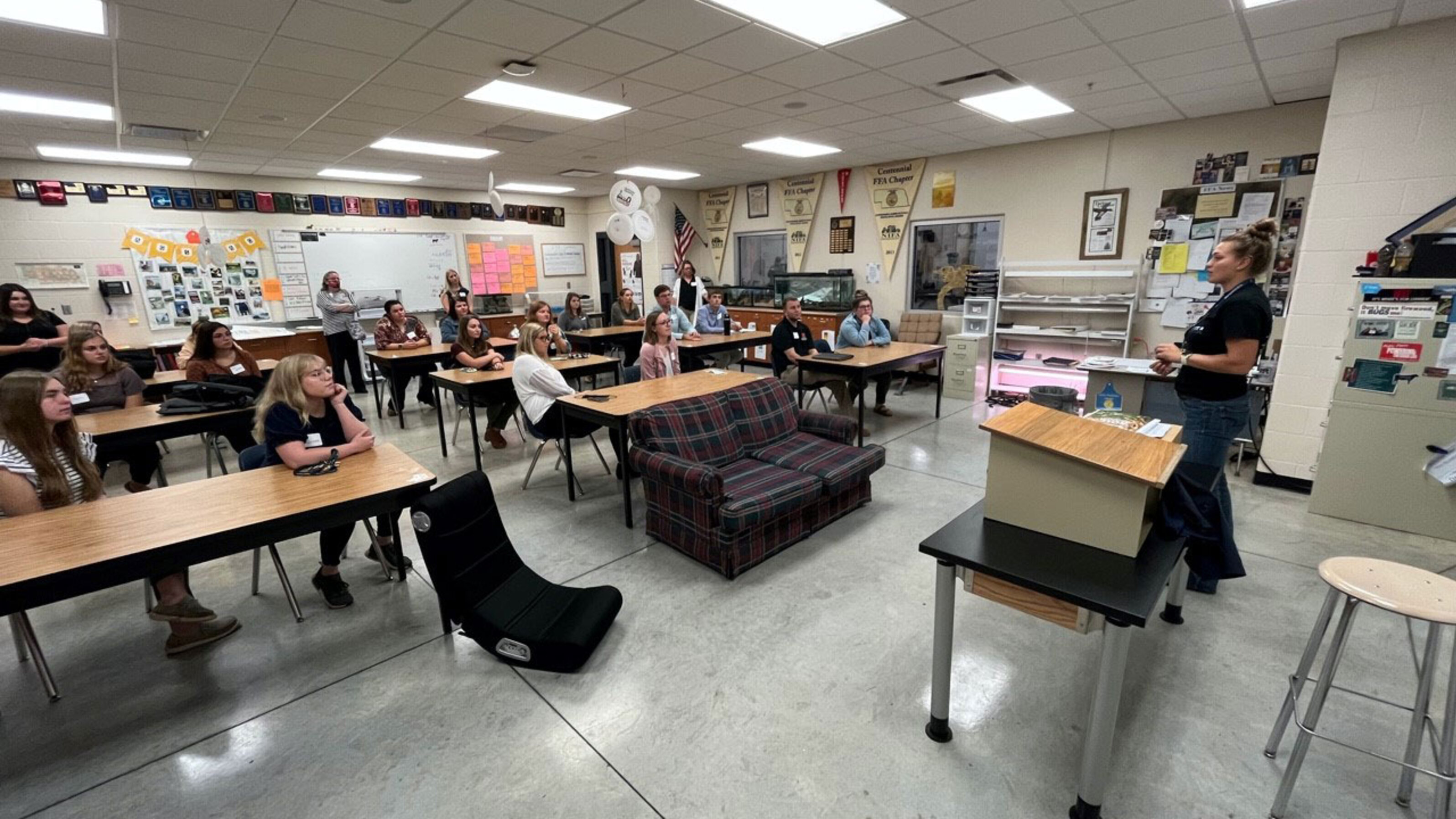 Classroom with students seated at desks, listening to a teacher standing near the front.